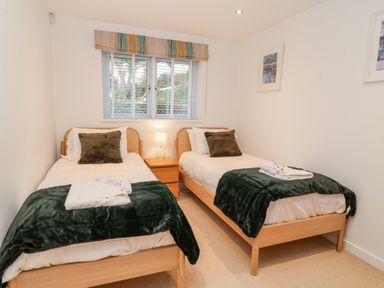 A bedroom with two single beds, bedside table with lamp, window with blinds and valance, and framed pictures on walls at A Stone's Throw in Downderry