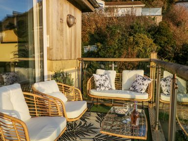 An outdoor balcony with cushioned chairs a loveseat glass table with wine glasses and a bottle at A Stone's Throw in Downderry