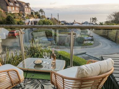A balcony with two chairs a glass table with two wine glasses and a bottle overlooking a street and the sea at A Stone's Throw in Downderry