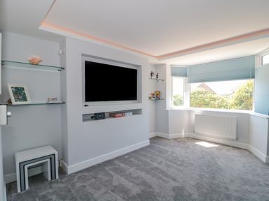 A living room with a mounted television shelves nested tables and a large window with blinds at Fisherman's Rest in Wyke Regis