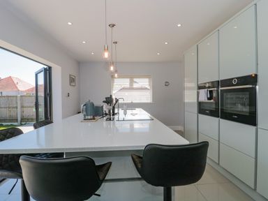 A kitchen with white cabinets a large island with black chairs and hanging light bulbs at Fisherman's Rest in Wyke Regis