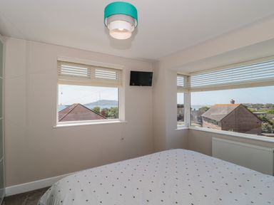 A bedroom with two windows showing a view of rooftops and hills with a bed and a small wall-mounted TV at Fisherman's Rest in Wyke Regis