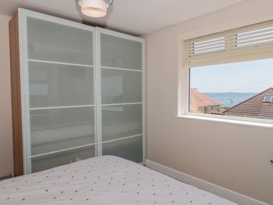 A bedroom with a bed a wardrobe with frosted glass doors and a window with a view of rooftops and the sea at Fisherman's Rest in Wyke Regis