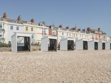 A row of white beach huts on a pebbled shore with houses in the background at Fisherman's Rest in Wyke Regis
