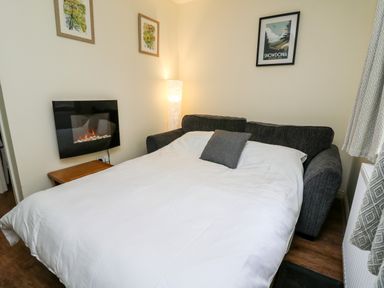 A sofa bed pulled out with white bedding and a grey pillow in a living room with wall art a floor lamp and a fireplace at Honeysuckle Cottage in Crossgates