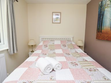 A bedroom with a bed covered with a patchwork quilt two rolled white towels on the bed two bedside tables with lamps one with painting on wall and window with curtains at Honeysuckle Cottage in Crossgates