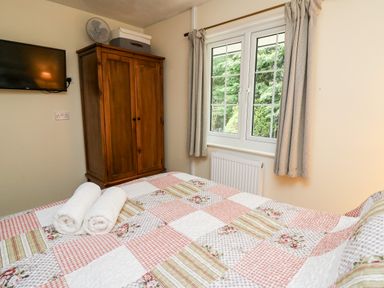 A bedroom with a patterned quilt bedspread rolled towels a wooden wardrobe a wall mounted television and a window with curtains at Honeysuckle Cottage in Crossgates