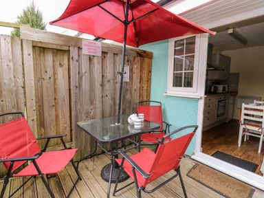 An outdoor patio with a glass table under a red umbrella and red chairs next to a wooden fence and an open door showing a kitchen at Honeysuckle Cottage in Crossgates