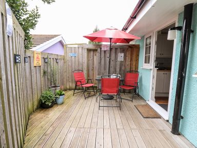 A wooden deck patio with red chairs and umbrella a table and potted plants outside a teal building at Honeysuckle Cottage in Crossgates