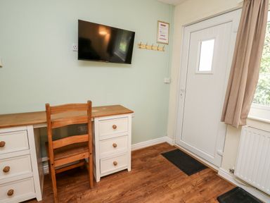 A wooden desk with drawers and a wooden chair under a wall-mounted television next to a white door with a window at Primrose Cottage in Crossgates