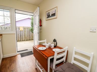 A small dining area with a wooden table set for three and white chairs next to an open door leading outside at Primrose Cottage in Crossgates