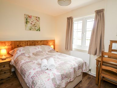A bedroom with a double bed with floral quilt and two rolled towels on it next to a wooden nightstand with lamp and a wooden dressing table with chair by a window at Primrose Cottage in Crossgates