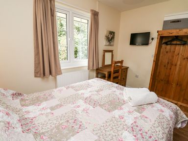 A bedroom with a floral quilted bedspread wooden dressing table chair wall-mounted tv and window with curtains at Primrose Cottage in Crossgates