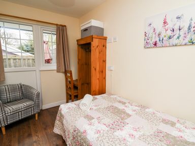 A bedroom with a single bed covered with a floral quilt a rolled towel on the bed a wooden wardrobe with a chair next to a window and a small armchair near the door at Primrose Cottage in Crossgates