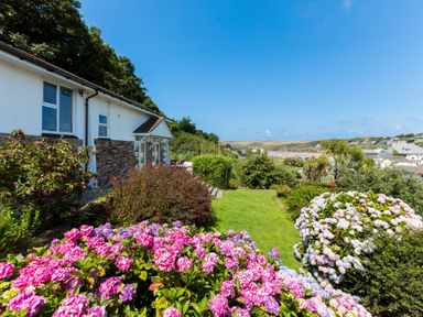 A garden with pink and white flowers next to a house with a view of a coastal town and hills at Beach Haven in Polzeath