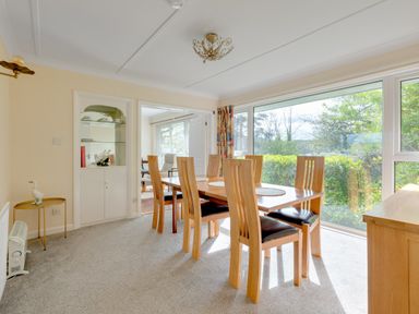 A dining room with a wooden table and six chairs next to large windows at Beach Haven in Polzeath