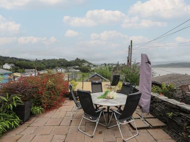 An outdoor patio with a round table and six chairs surrounded by plants overlooking a village and water at Alltwen in Borth-y-Gest