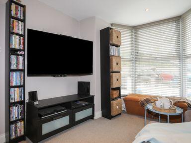 A living room with a wall-mounted television a media console bookshelf with DVDs a tall shelving unit with wicker baskets an ottoman by windows with blinds and a small table with a tea set at Alltwen in Borth-y-Gest
