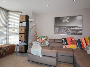 A living room with a gray sectional sofa colorful pillows a tall shelving unit with baskets a standing lamp a side table and a large black and white photograph on the wall at Alltwen in Borth-y-Gest