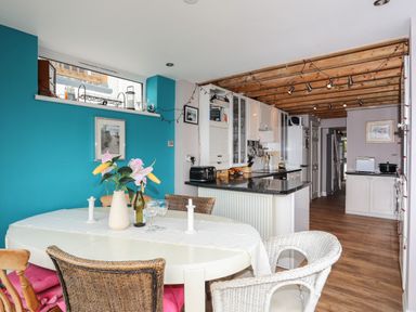A dining area with a round table and various chairs next to a kitchen with white cabinets and wooden ceiling beams at Alltwen in Borth-y-Gest