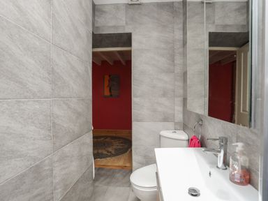 A bathroom with gray tiled walls a white sink and toilet and a doorway leading to a room with a red wall at Alltwen in Borth-y-Gest