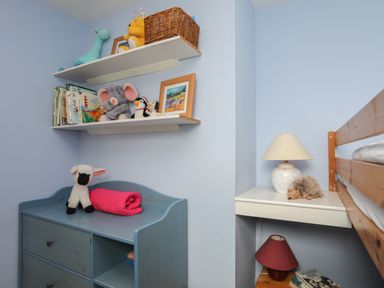 A corner of a blue painted room with shelves holding stuffed animals and books a blue dresser with a stuffed lamb and pink blanket a bedside table with two lamps and books a wooden bunk bed at Alltwen in Borth-y-Gest