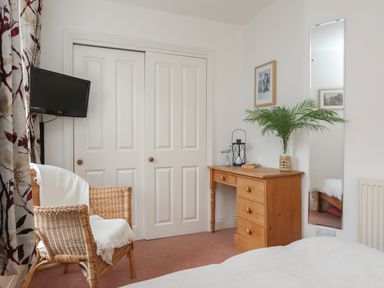 A bedroom with a wicker chair draped with a white cloth a wooden dresser with a plant and decor a wall mirror a mounted TV and white closet doors at Alltwen in Borth-y-Gest