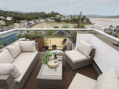 An outdoor seating area with cushioned wicker furniture and a glass table with a bottle of wine overlooking a coastal village and beach at Alltwen in Borth-y-Gest