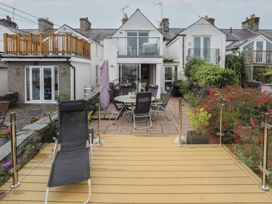 A patio with a round table and chairs and a deck with a lounge chair outside a row of white houses at Alltwen in Borth-y-Gest