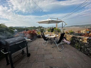 A patio with a barbecue grill table chairs and an umbrella overlooking a hilly town and water at Alltwen in Borth-y-Gest