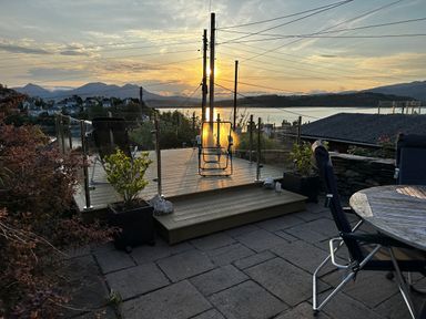 A patio with outdoor chairs and a round wooden table overlooking a lake and mountains at sunset at Alltwen in Borth-y-Gest