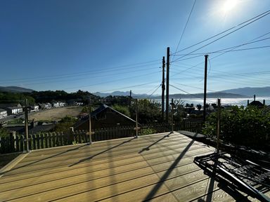 A wooden deck with glass railings overlooking a village with houses and hills in the background at Alltwen in Borth-y-Gest