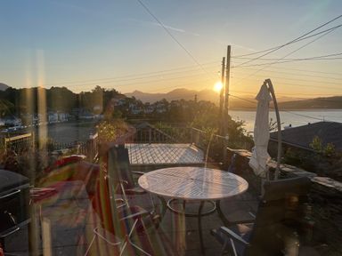 A patio with a round table chairs and a folded umbrella overlooking a waterfront village at sunset at Alltwen in Borth-y-Gest