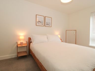 A bedroom with a wooden bed with white bedding bedside tables with lamps two framed pictures on the wall and a wooden clothes rack by the window at Apartment 1 in Exmouth