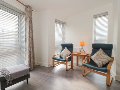 A sitting area with two armchairs a wooden side table with a lamp and large windows with blinds and curtains at Apartment 9 in Exmouth