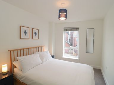 A bedroom with a wooden bed frame white bedding two framed pictures a window and a lamp at Apartment 9 in Exmouth