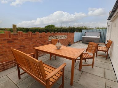 An outdoor patio with wooden table and chairs a hot tub and a brick wall at Bryn Eglwys Bach in Penmynydd near Llanfairpwllgwyngyll