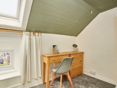 A small wooden desk with a mirror and a gray chair under a slanted green ceiling next to a window with white curtains at Stonewall Sandsend