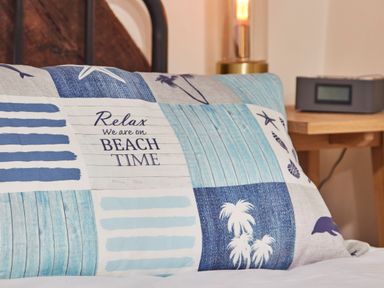 A pillow with blue and white beach-themed patterns on a bed near a wooden nightstand with a lamp and a clock