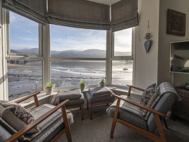 A sitting area with two armchairs, two ottomans, small plants on the window sill, and a view of the water and hills at Harbour View - Flat 2 in Barmouth
