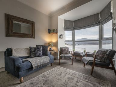 A sitting room with a sofa chairs by a large window overlooking water and a bridge at Harbour View - Flat 2 in Barmouth