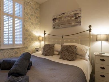 A bedroom with a double bed two bedside tables with lamps and a window with white shutters at Harbour View - Flat 2 in Barmouth
