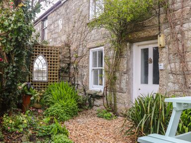 An outdoor stone wall with a white door and window surrounded by plants and a mirror on a lattice with a light blue bench at Rose Cottage in Lower Bostraze near St Just