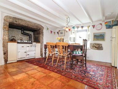 A dining area with a wooden table and chairs on a patterned rug next to a stove set in a stone alcove at Rose Cottage in Lower Bostraze near St Just