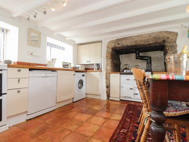 A kitchen with white cabinets dishwasher washing machine stove under a large stone fireplace and a dining table with wooden chairs at Rose Cottage in Lower Bostraze near St Just