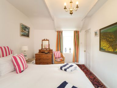A bedroom with a bed with striped pillows a wooden dresser an armchair and colorful curtains at Rose Cottage in Lower Bostraze near St Just