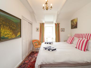 A bedroom with a double bed with striped pillows and towels on it a window with striped curtains a chair a painting and a chandelier at Rose Cottage in Lower Bostraze near St Just