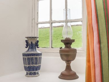 A blue and white vase and an oil lamp on a windowsill with a green field outside at Rose Cottage in Lower Bostraze near St Just