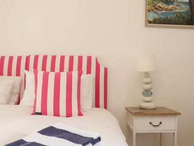 A bedroom with a bed having pink and white striped cushions and headboard and a wooden bedside table with a lamp at Rose Cottage in Lower Bostraze near St Just