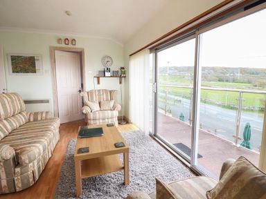 A sitting room with striped sofas a wooden coffee table a wall clock and large sliding glass doors overlooking a road and fields at Morlyn Guest House Apartment in Llandanwg near Harlech